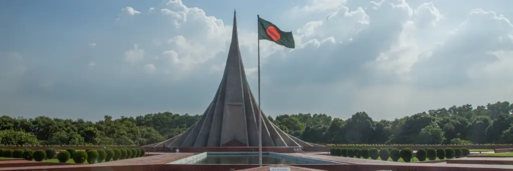 Monument with tall, pointed concrete spire behind a flagpole flying the Bangladesh flag, reflected pool and manicured shrubs in the foreground under a partly cloudy sky.