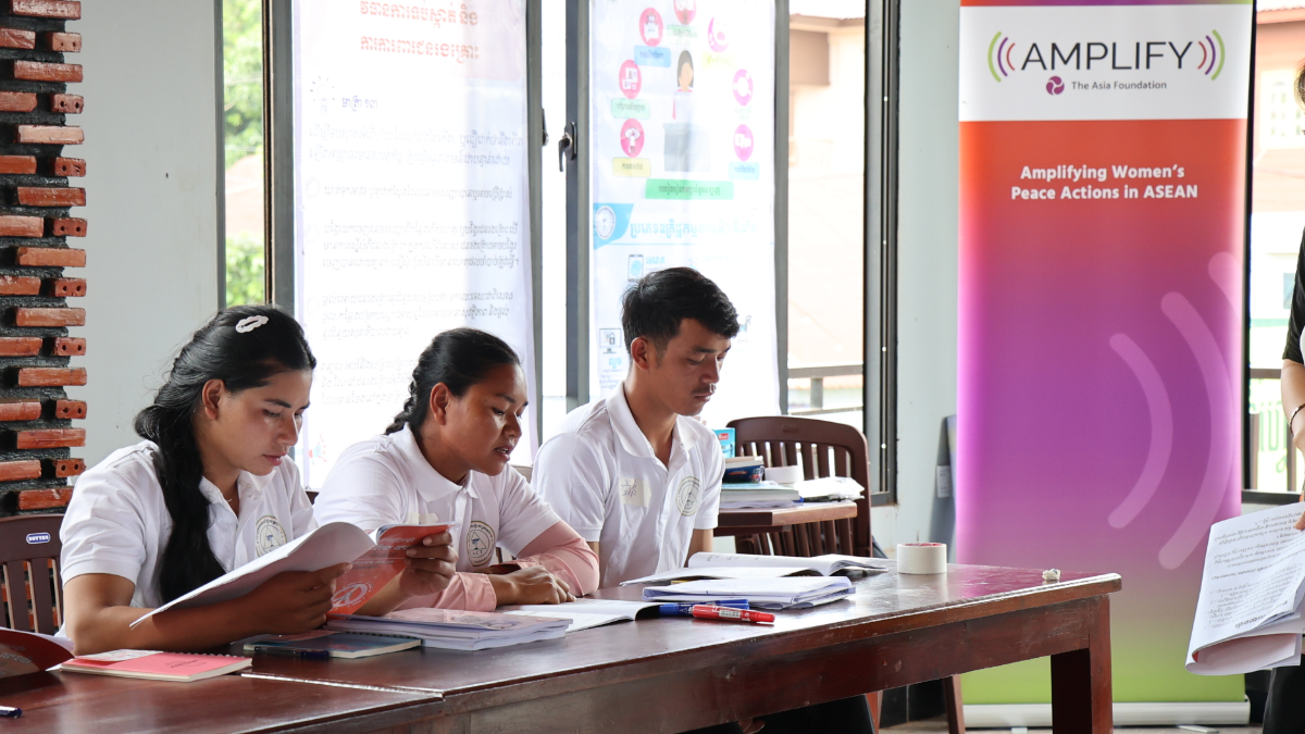 Three young adults in white polo shirts sit at a wooden table reading documents in a classroom; an AMPLIFY banner reading “Amplifying Women’s Peace Actions in ASEAN” stands to the right.