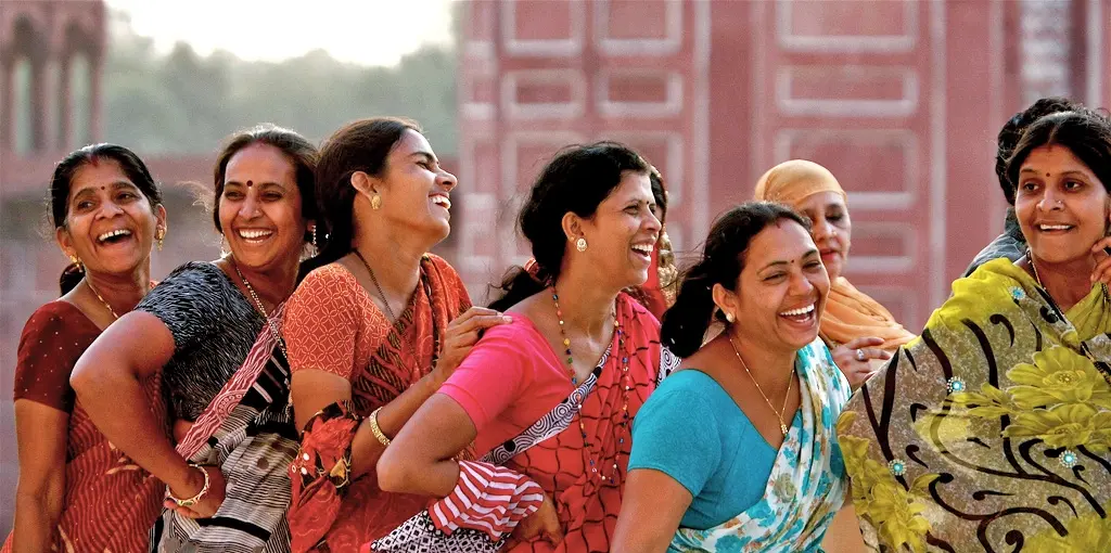 A row of smiling women in colorful saris, linked arm-in-arm and laughing outdoors before red-brick buildings.