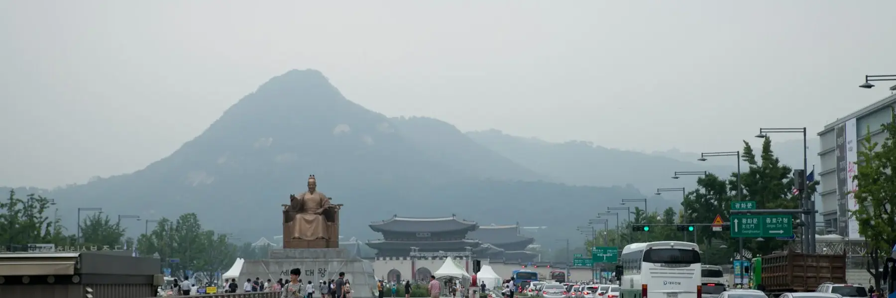 Wide pedestrian plaza in downtown Seoul with the bronze statue of King Sejong seated on a pedestal in front of Gwanghwamun gate; hazy mountain rises in the background, people walk the plaza and cars line the road to the right.