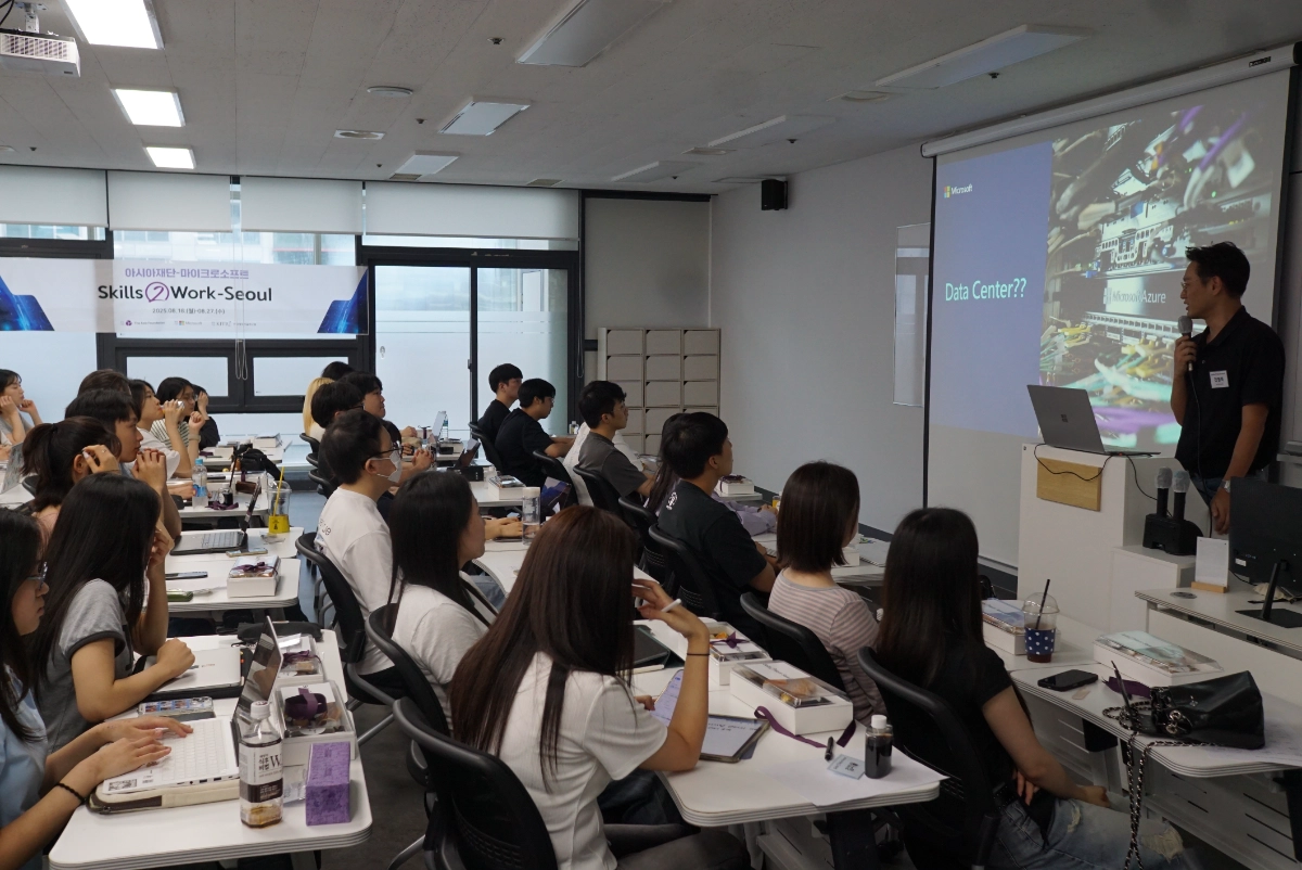 A classroom-style workshop with a presenter speaking into a microphone at a laptop and projecting a slide titled “Data Center??” while about 20 students sit at desks with laptops and notebooks; a banner at the back reads “Skills2Work–Seoul.”