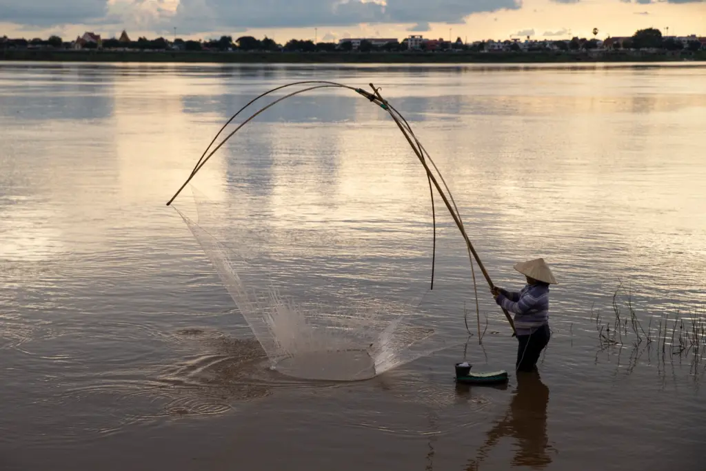 Person wearing a conical hat and striped sweater stands waist-deep in a wide river at sunset, holding long bamboo poles to cast a large circular fishing net that fans out over the water; distant shoreline and soft clouded sky form a low silhouette on the horizon.