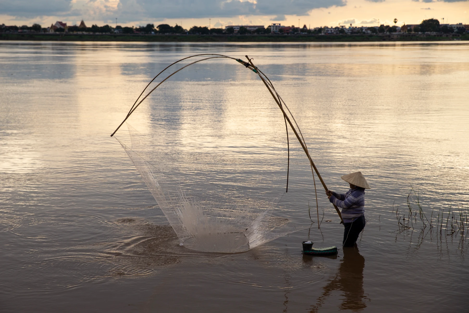Person wearing a conical hat and striped sweater stands waist-deep in a wide river at sunset, holding long bamboo poles to cast a large circular fishing net that fans out over the water; distant shoreline and soft clouded sky form a low silhouette on the horizon.