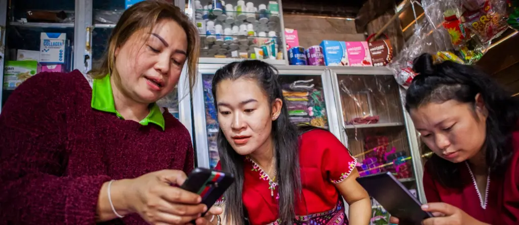 Three women in a small shop leaning over phones, surrounded by colorful woven textiles and shelves.