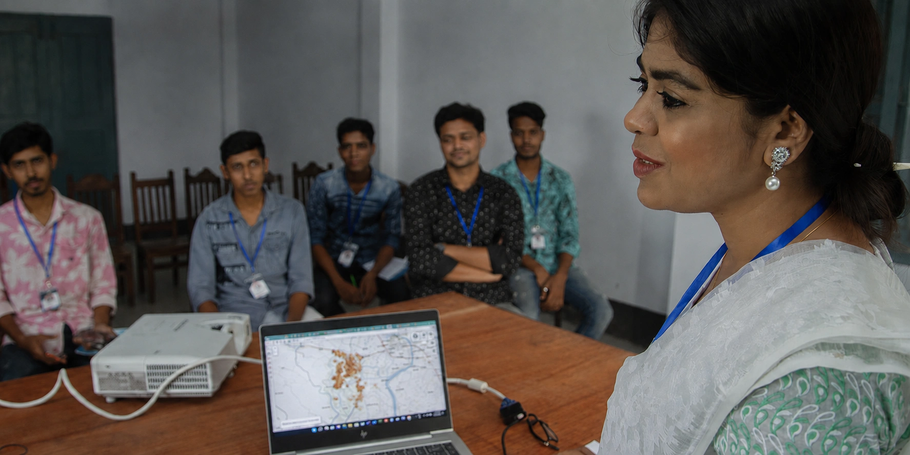 Woman speaking at a table to five seated students; a laptop showing a map and a projector sit on the table in a classroom.