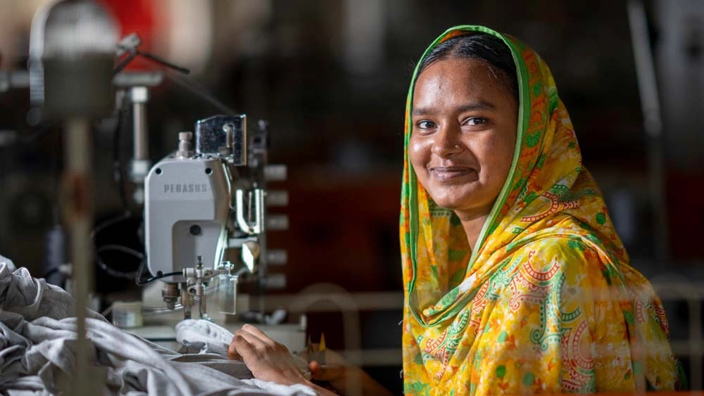 A sewing machine operator, Tazmin Akhter, smiles as she works her shift. Her precise stitching and attention to detail ensure that every garment meets the highest quality standards. Fakir Apparels Ltd. Narayanganj, Dhaka.