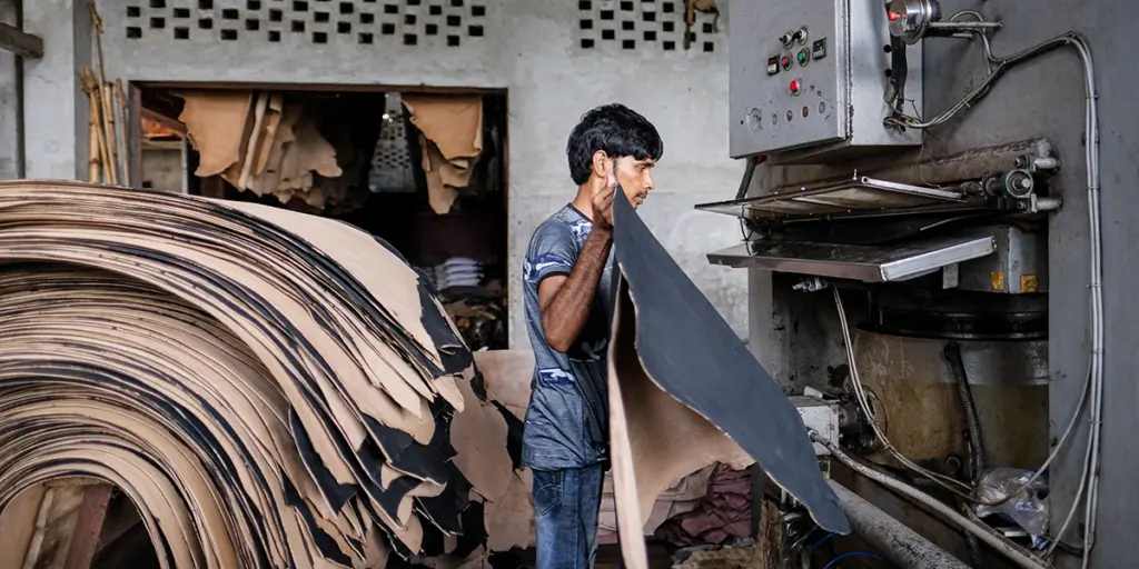 Worker in a tannery feeding a large sheet of leather into an industrial press, with stacked hides piled to the left and machinery on the right.