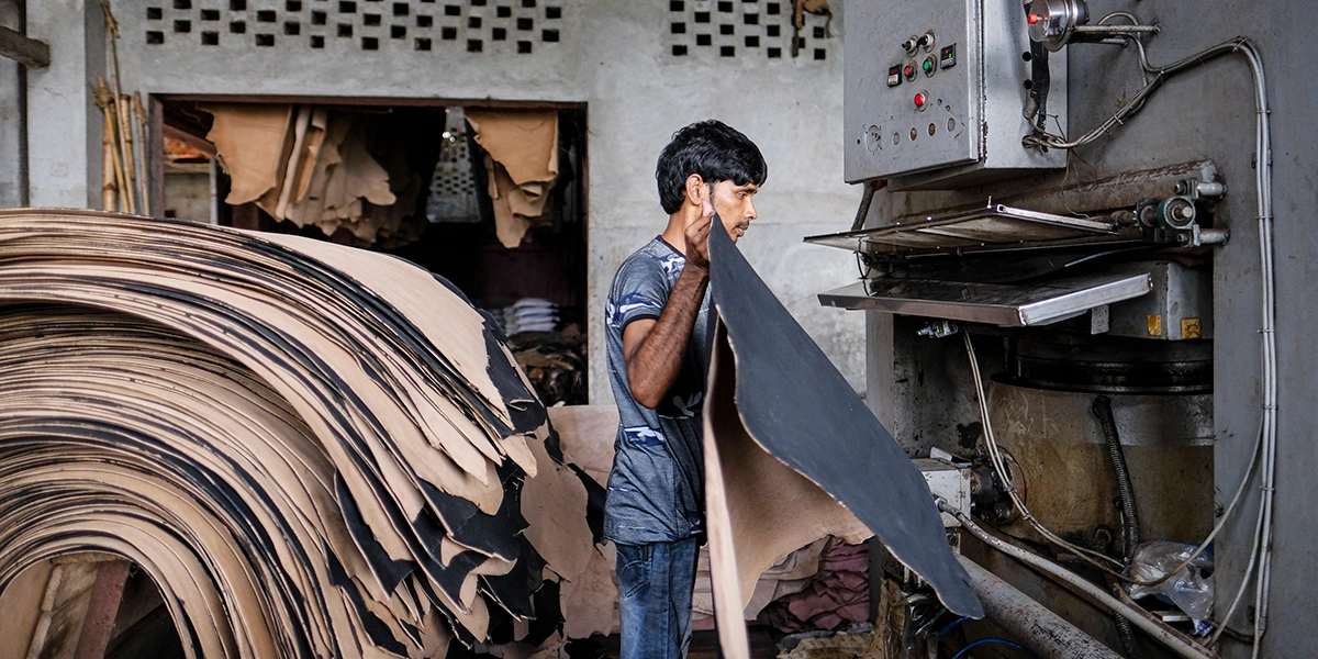 Worker in a tannery feeding a large sheet of leather into an industrial press, with stacked hides piled to the left and machinery on the right.