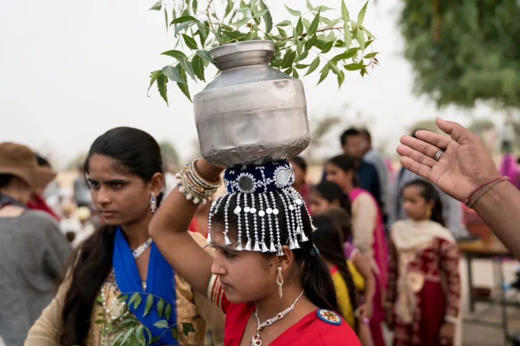A young Rajasthani woman in traditional dress balancing a metal kalash (pot) adorned with neem leaves on her head during a ceremonial procession in Rajasthan, India.