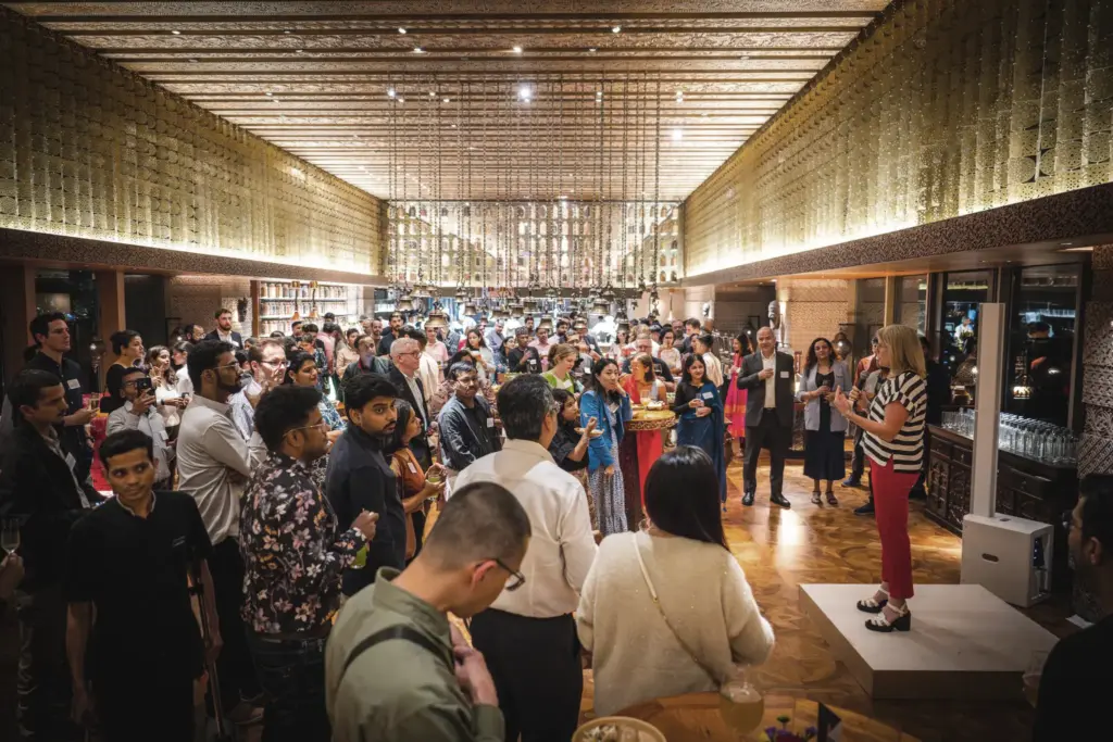 A wide-angle photo of a woman in a striped shirt and red pants speaking on a microphone to a large audience of business professionals gathered for an event.