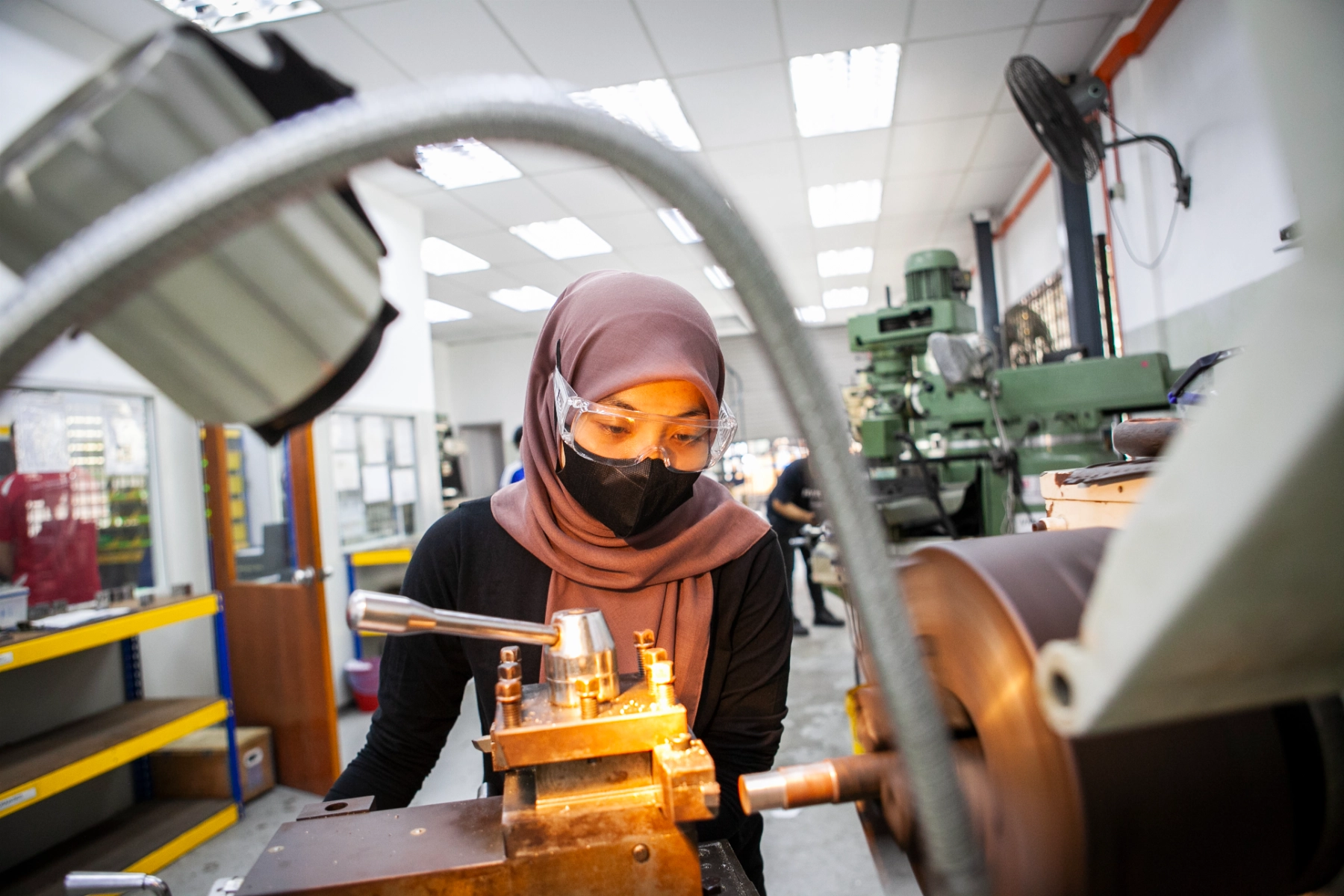 A young woman works safely and confidently at a lathe in a technical workshop in Malaysia, wearing safety glasses and a mask as she operates precision machinery.