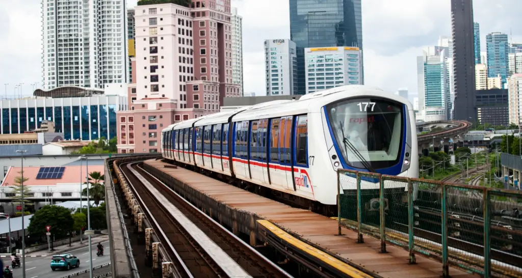 A Rapid KL LRT train (unit 177) travels along an elevated track through Kuala Lumpur, with a dense city skyline of high-rise towers in the background under an overcast sky.